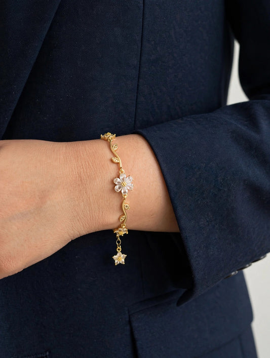 A delicate gold chain bracelet with a flower and star charm, shown worn on a person's wrist against a dark navy blazer.