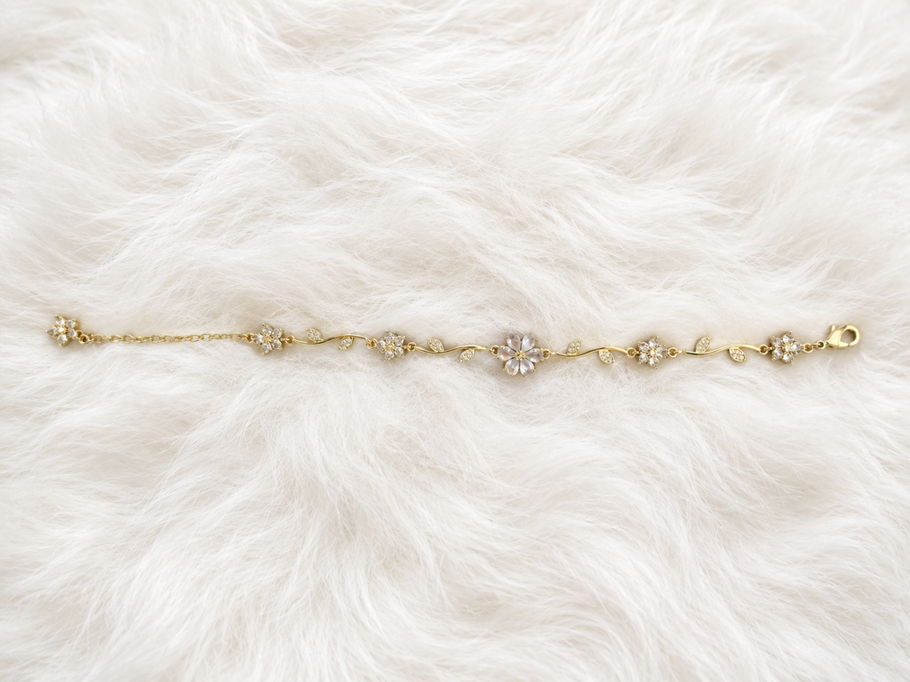 A delicate gold chain bracelet with five floral clusters, featuring clear crystals in the center of each flower, laid flat on a white faux fur background.