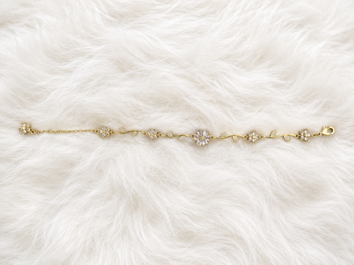 A delicate gold chain bracelet with five floral clusters, featuring clear crystals in the center of each flower, laid flat on a white faux fur background.