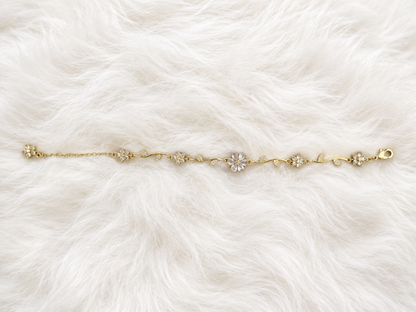 A delicate gold chain bracelet with five floral clusters, featuring clear crystals in the center of each flower, laid flat on a white faux fur background.